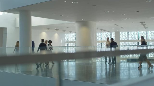Slow motion shot of businessmen and businesswomen walking through corridor of modern office building with glass walls; two colleagues standing and discussing something