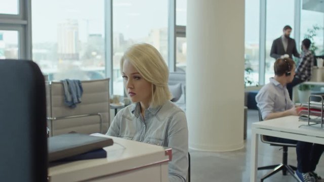 Tracking shot of young blonde woman typing on computer; office workers collaborating in modern workspace; two business partners discussing something in the background