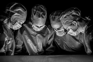 Black white photo of doctors in the operating room. Doctors are dressed in surgical suits, their faces have medical masks, and on their heads are surgical caps.