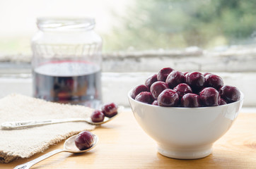 Cherries from compote in a white ceramic bowl, two silver spoons and a jar with cherry juice in the background