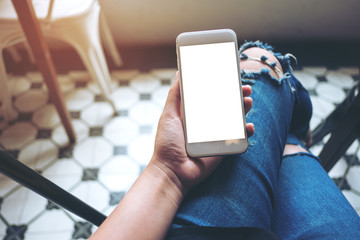 Mockup image of hand holding white mobile phone with blank white screen on thigh with a vintage tile floor in cafe , feeling relaxed