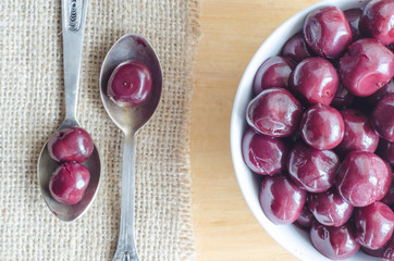 Cherry compote in a white ceramic bowl and two silver spoon on a wooden board - top view