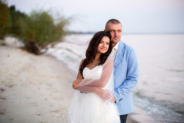Portrait of Couple of lovers newlyweds on the seafront. Brunette bride in wedding dress with a beautiful hairstyle, veil and makeup and her husband