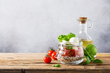 Delicious caprese salad with quinoa, ripe cherry tomatoes and mini mozzarella cheese with fresh basil leaves in glass jar and olive oil. Italian healthy food concept with copy space.