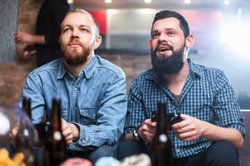Men with a beard sitting on the couch at home with beer and chips with joysticks in hand playing computer video games. The concept of friendship, technology and weekend
