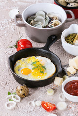 Breakfast with fried quail eggs in iron cast pan, cherry tomatoes, onion, ketchup sauce, seasonings in mortar, cheese grater, egg shell in pot. Beige concrete texture background. Close up.