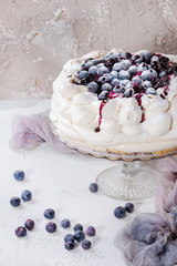 Homemade meringue cake Pavlova with whipped cream, sugar powder, fresh blueberries and blueberry sauce on vintage cake stand on beige concrete texture background, gauze textile.