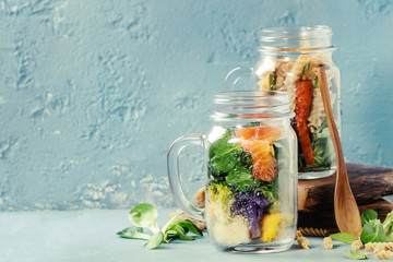 Variety of vegetable salads in mason jars. Salad with greens, pasta, carrots, cauliflower, salmon. Standing with wooden spoon and serving board over blue texture background. Healthy food to go.