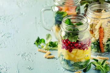 Variety of salads in mason jars. Fruit salad mango, pomegranate, greens, vegetables, wholegrain pasta, carrots and cauliflower, salmon. Standing over blue texture background. Food to go.