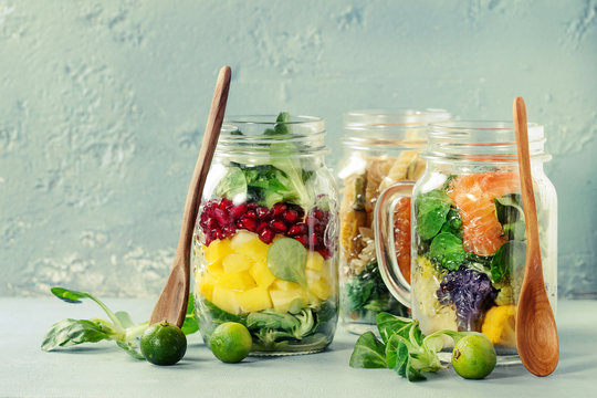 Variety Of Salads In Mason Jars. Fruit Salad Mango, Pomegranate, Greens, Vegetables Pasta, Carrots, Cauliflower, Salmon. Standing With Mini Limes And Wood Spoon Over Blue Background. Food To Go.