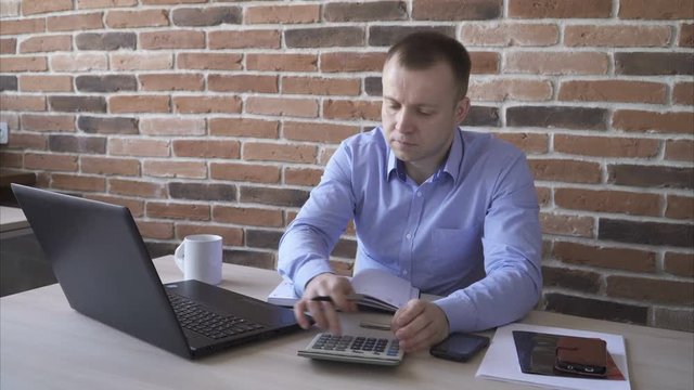 Man accountant at work, counting using a calculator and a laptop, brick wall background