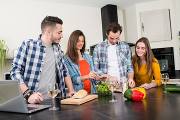 group of friends, four people man and woman at home cooking, drinking wine and preparing food meal together in the kitchen