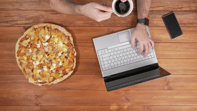 A man checks the messages on the phone and eats pizza. Nearby there is a coffee mug. Wooden table top view