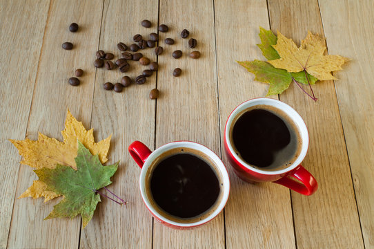 Two cups of coffee with autumn leaves and coffee beans on a wooden background