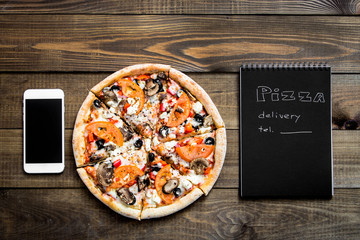 Close-up of pizza, a mobile phone and a notebook with the text: Pizza delivery. background wooden table. Notebook black with white text