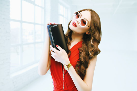 Indoor portrait of young beautiful fashionable woman posing in white interior. Model wearing stylish round sunglasses, red clothes, holding small leather clatch bag. Female fashion concept. Waist up