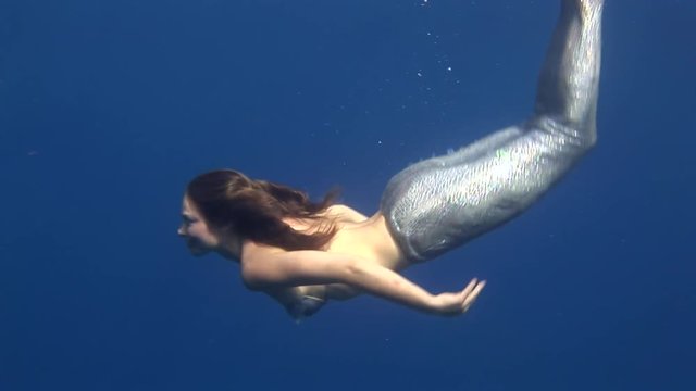 Young girl model underwater nixie costume on blue background poses in Red Sea. Filming a movie at camera. Extreme sport in marine landscape, coral reefs, ocean inhabitants.