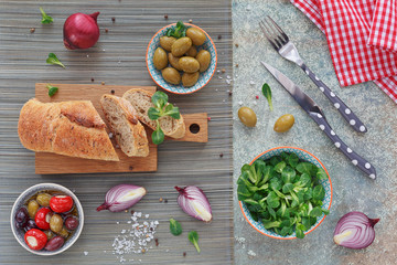 Green and black olives, loaf of fresh multigrain bread, corn salad and red onion over old wood background.