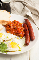 Fried eggs, beans, grilled sausages, bread on a plate on a white wooden background.
