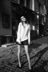 Pretty girl wearing heels standing in the street. Fashion model in the city next to a cafe. Skinny teenager with a long hair in a hat street style. Black and white fashion photography.