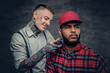 Obraz premium Tattooed barber cutting the beard of a Black hipster male.