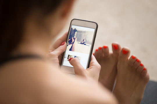Young attractive woman sitting and checking pictures after yoga photo session on her smartphone, enjoying and posting good shots, using filter application for images, view over the shoulder, closeup