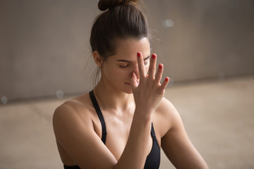 Young attractive yogi woman practicing yoga, using Alternate Nostril Breathing, making nadi shodhana pranayama therapeutic exercise, working out, cool urban style grey studio background, closeup,