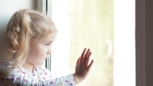  Little girl stares sadly out of a  window. Sitting on the window sill. The child looks out of the window.