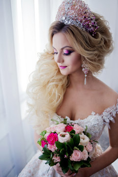 Young fashion bride with perfect skin and make up, curly hair, flowers and tiara on the head, against a window indoors