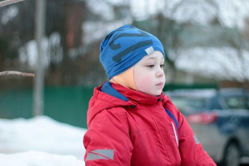 Very sad cute little boy sits on top of a snowy hill in bright clothes.