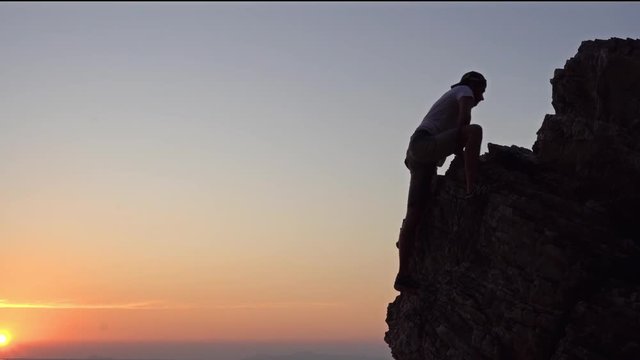 Young man climbs on the rock against colourful sunset background