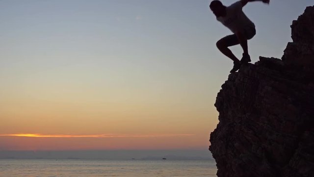 Young man jumping from the cliff against colourful sunset background