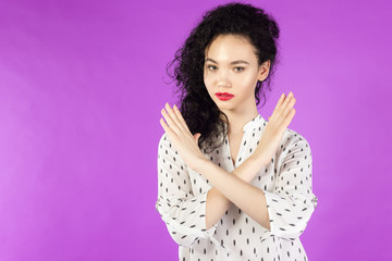 young curly brunette woman in a shirt says no hands crossed on a pink background