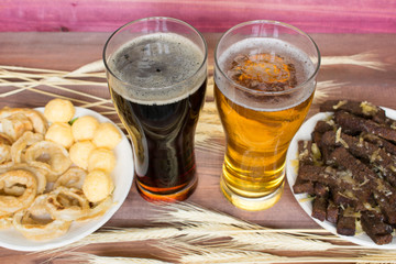 Two glasses of beer with snacks and wheat on the brown wooden background.