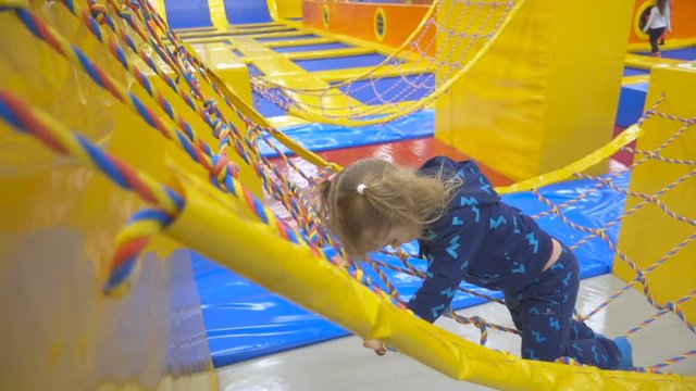 Girl is Walking by the Bridge Through rope Stairs Little Girl with Long Fair Hair in Indoors Playground. Active Toddler Girl Having Fun At Sport Center. Slow Motion