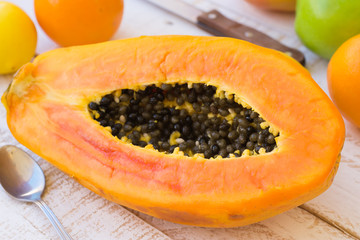 Ripe juicy halved papaya on kitchen table with citrus fruits and apples, knife on plank wood table, top view