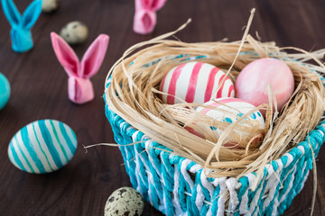 Colorful Easter Eggs on the rustic dark wooden background with nest, blue basket, rabbit from a napkin. Selective focus.