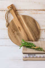 Round wood cutting board, stack of linen towels, rosemary twigs on white plank wood background, rural kitchen interior