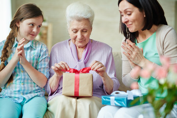 Girl and her mother clapping hands while aged woman opening gift-box