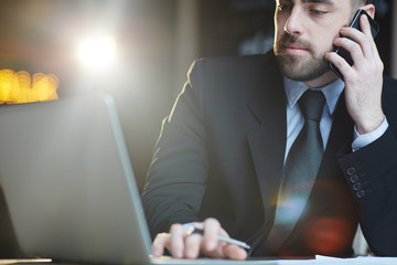 Portrait of businessman working in office using laptop and talking by mobile phone against black background with lens flare