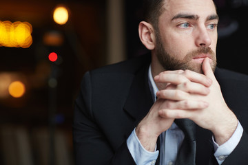 Portrait of modern bearded businessman looking away thinking about something resting head on clasped hands, against black background
