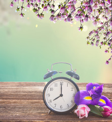 Spring time concept - retro alarm clock with flowers on wooden shelf, spring garden in background