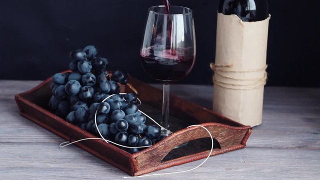 Pouring red wine into glass, Bunch - cluster of purple red grapes on wooden tray. Black background, wooden table.