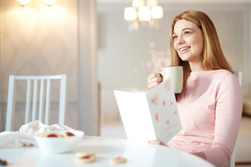 Young woman with greeting-card and drink sitting by table in the kitchen © pressmaster