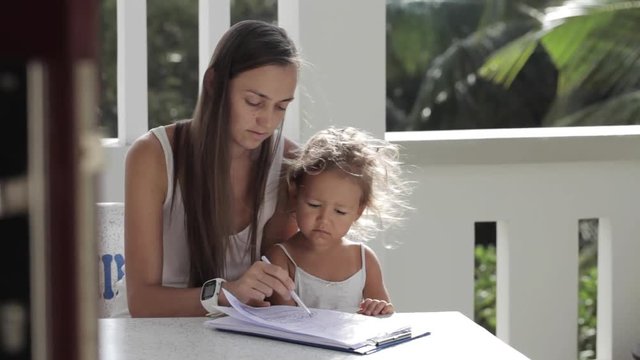 Young mother with daughter drawing at balcony with palm trees on the background