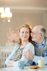 Portrait of smiling mature couple home at kitchen table: elderly man embracing elegant woman caringly and whispering compliments to her ear 