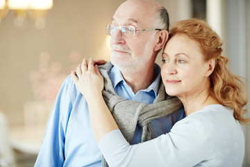 Portrait of loving elderly couple embracing and smiling looking away to window enjoying quiet retirement days at home