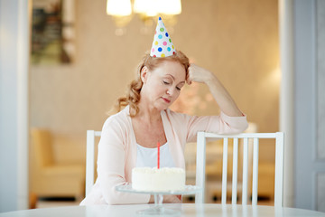 Portrait of sad  lonely mature woman sitting alone at birthday table with cake, wearing party hat and waiting for guests