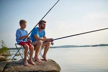Crédence de cuisine en verre imprimé Pêcher Portrait d& 39 un beau père barbu pêchant avec son fils assis sur un rocher dans un lac immobile avec des cannes à pêche et des engins aux beaux jours d& 39 été