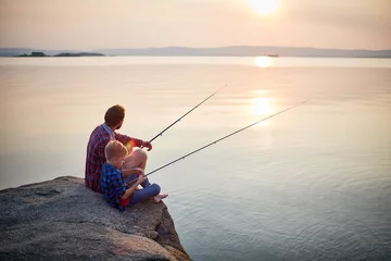 Fotobehang Vissen Achteraanzicht portret van vader en zoon samen zittend op rotsen vissen met hengels in kalme meerwateren met landschap van ondergaande zon